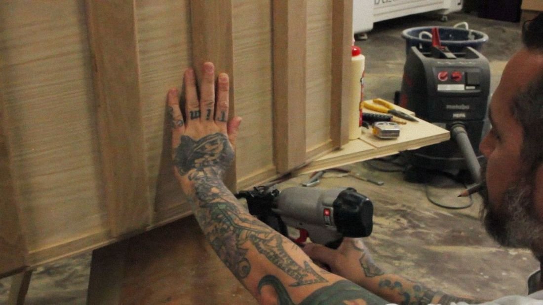 Tattooed hands holding a board steady while a nail gun fires, in a woodshop